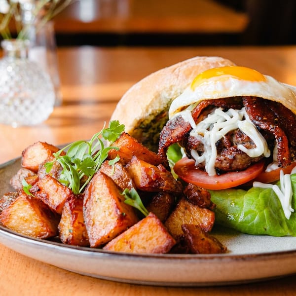 Burger with fried egg, bacon, cheese, lettuce, tomato, and roasted potatoes on a plate.