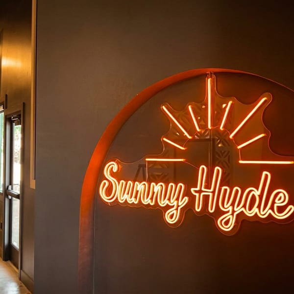 Bright orange 'Sunny Hyde Up' neon sign illuminates a modern restaurant interior with dining tables.