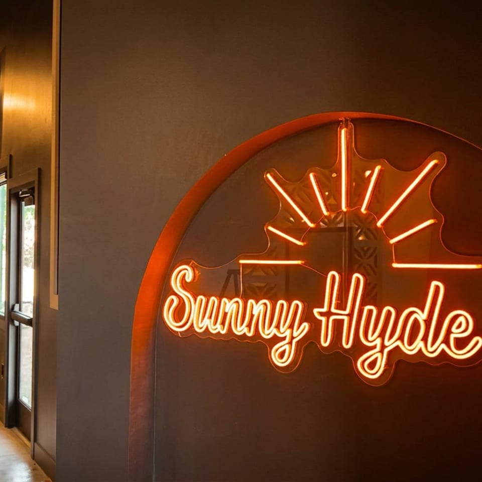 Bright orange 'Sunny Hyde Up' neon sign illuminates a modern restaurant interior with dining tables.
