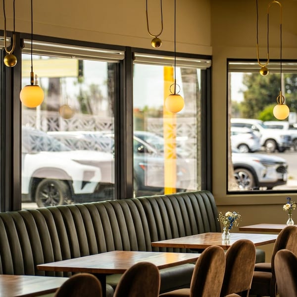 Modern restaurant interior with a long dark booth, wooden tables, and large windows overlooking a street.