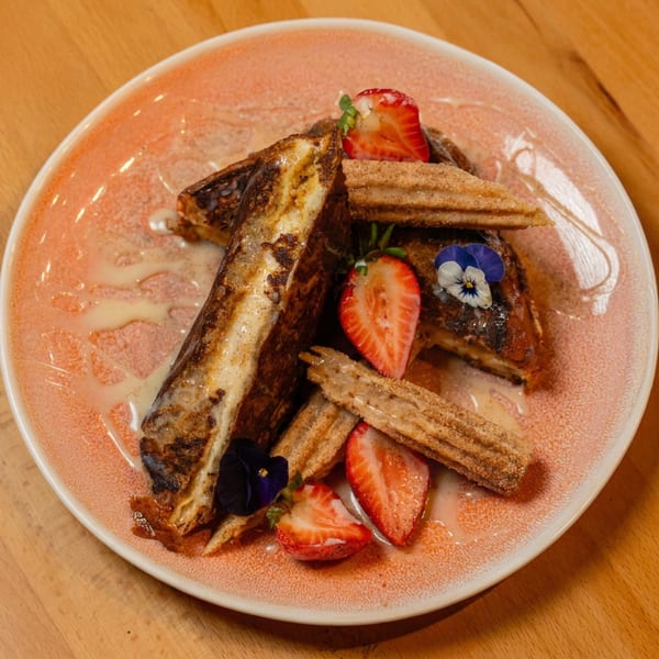 French toast and churros drizzled with sauce, garnished with strawberries and edible flowers on a pink plate.